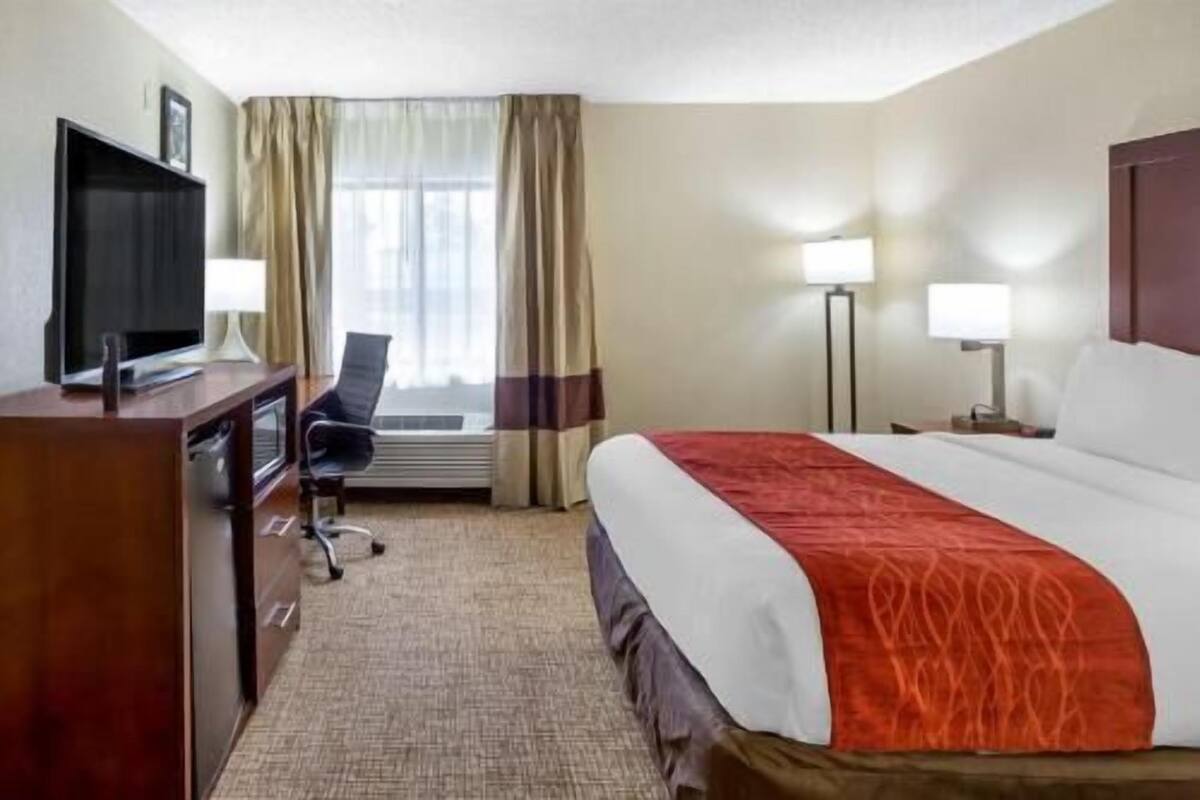 Habitación de hotel con cama, escritorio con televisor, silla, dos lámparas y ventana con cortinas.