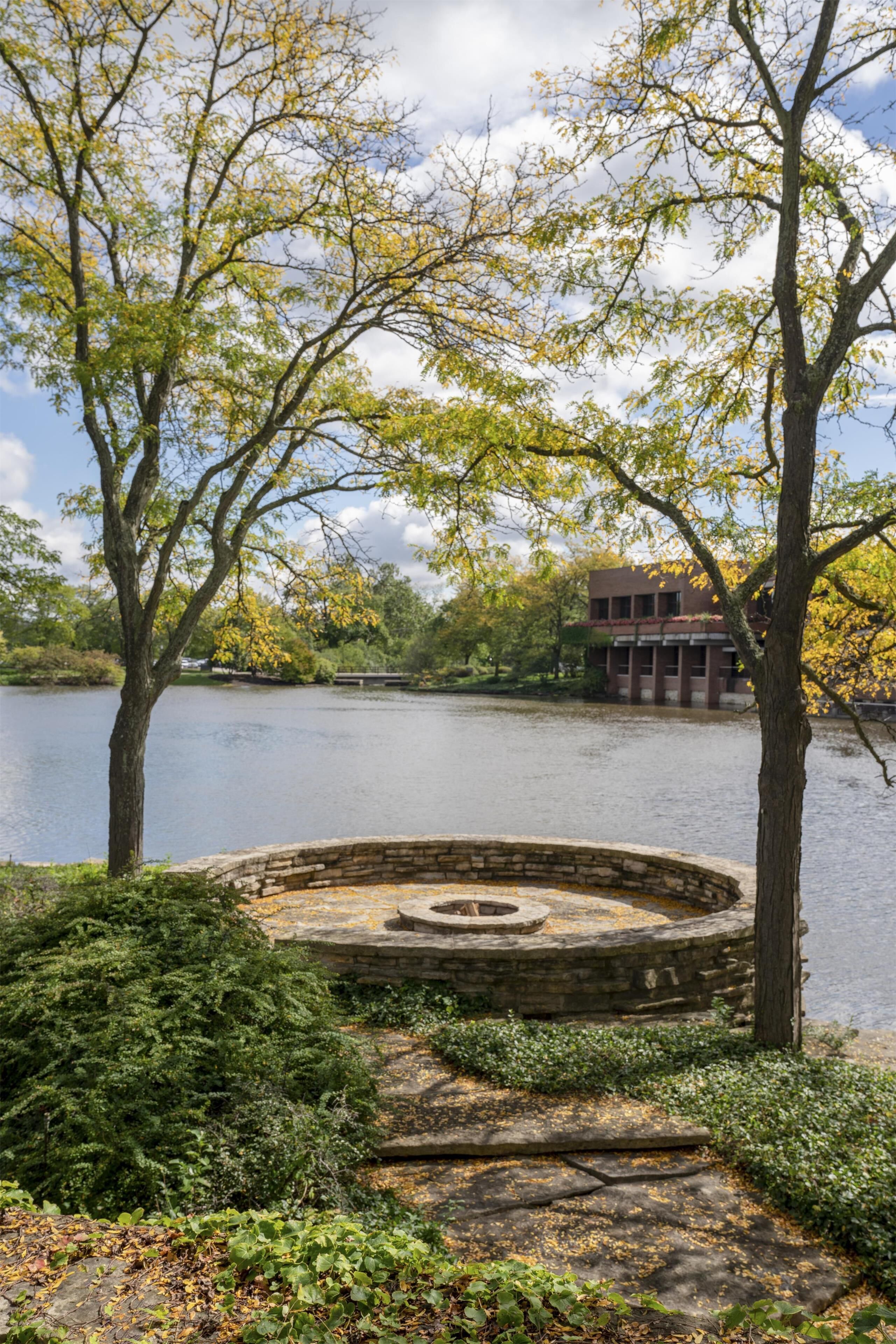 A stone fire pit surrounded by greenery, a body of water, and a building in the background.