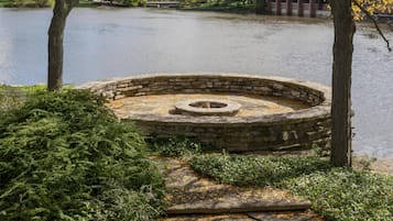 A stone fire pit surrounded by greenery, a body of water, and a building in the background.