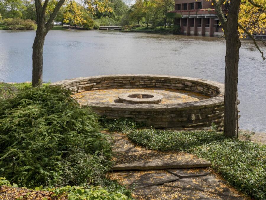 A stone fire pit surrounded by greenery, a body of water, and a building in the background.
