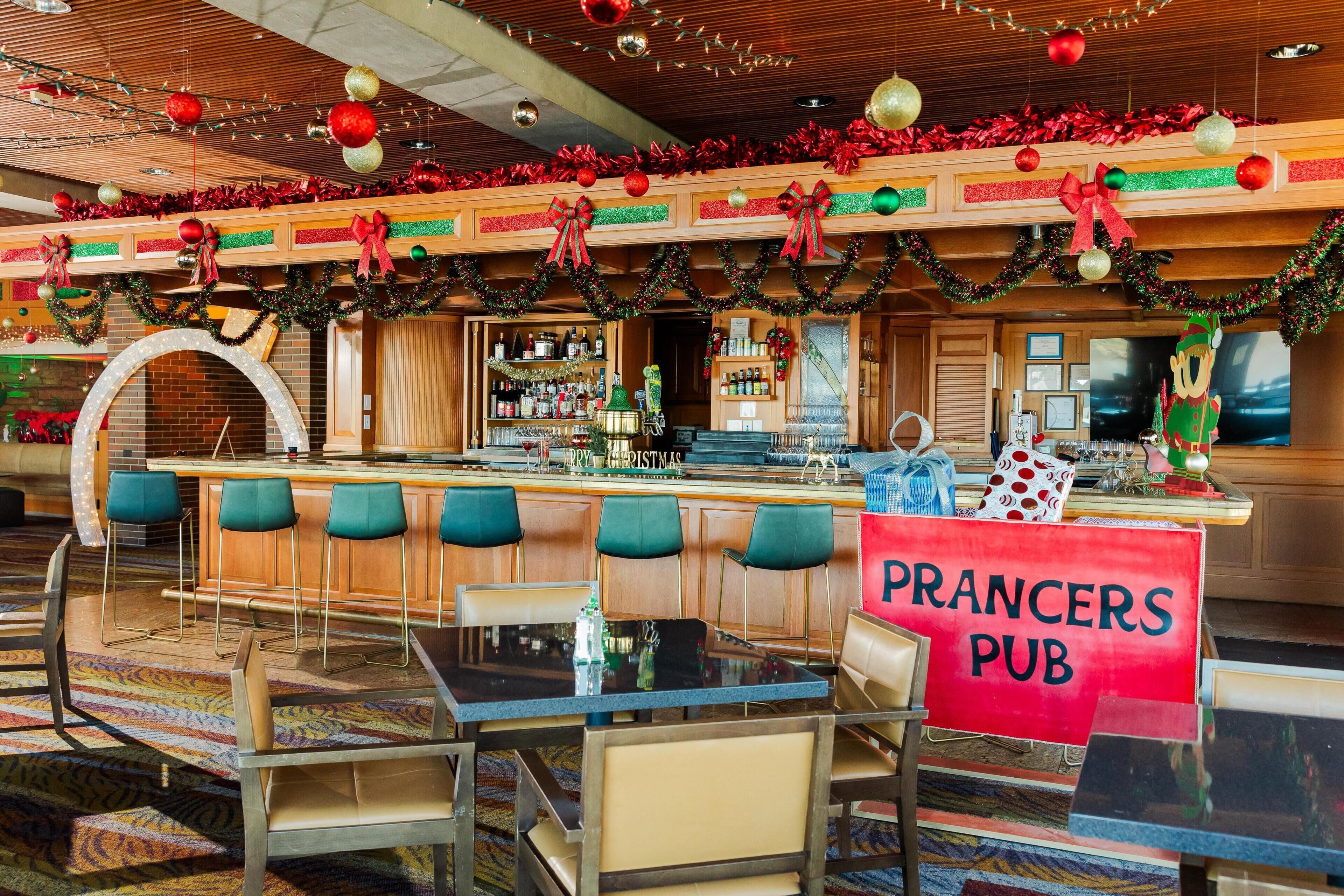 Un espace bar avec une enseigne indiquant « PRANCERS PUB », des tabourets de bar, un comptoir avec des bouteilles et une décoration de Noël festive.