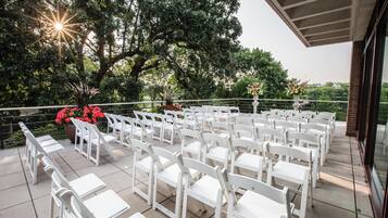 Un área de estar al aire libre con sillas y mesas blancas dispuestas en filas, rodeada de vegetación y un árbol grande.
