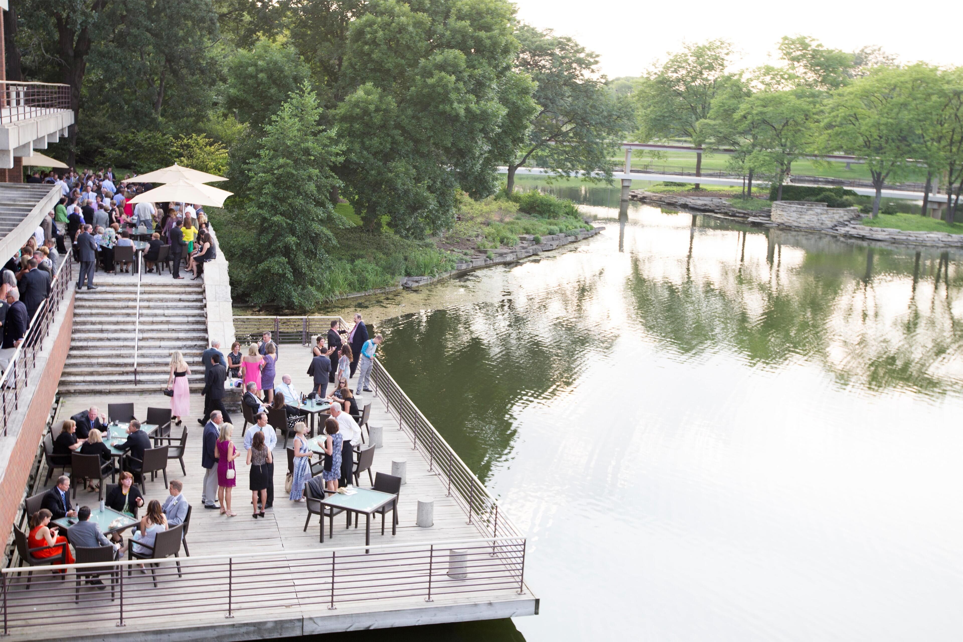 A riverside outdoor dining area with tables and chairs, surrounded by trees and a calm water body.