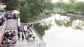 A riverside outdoor dining area with tables and chairs, surrounded by trees and a calm water body.