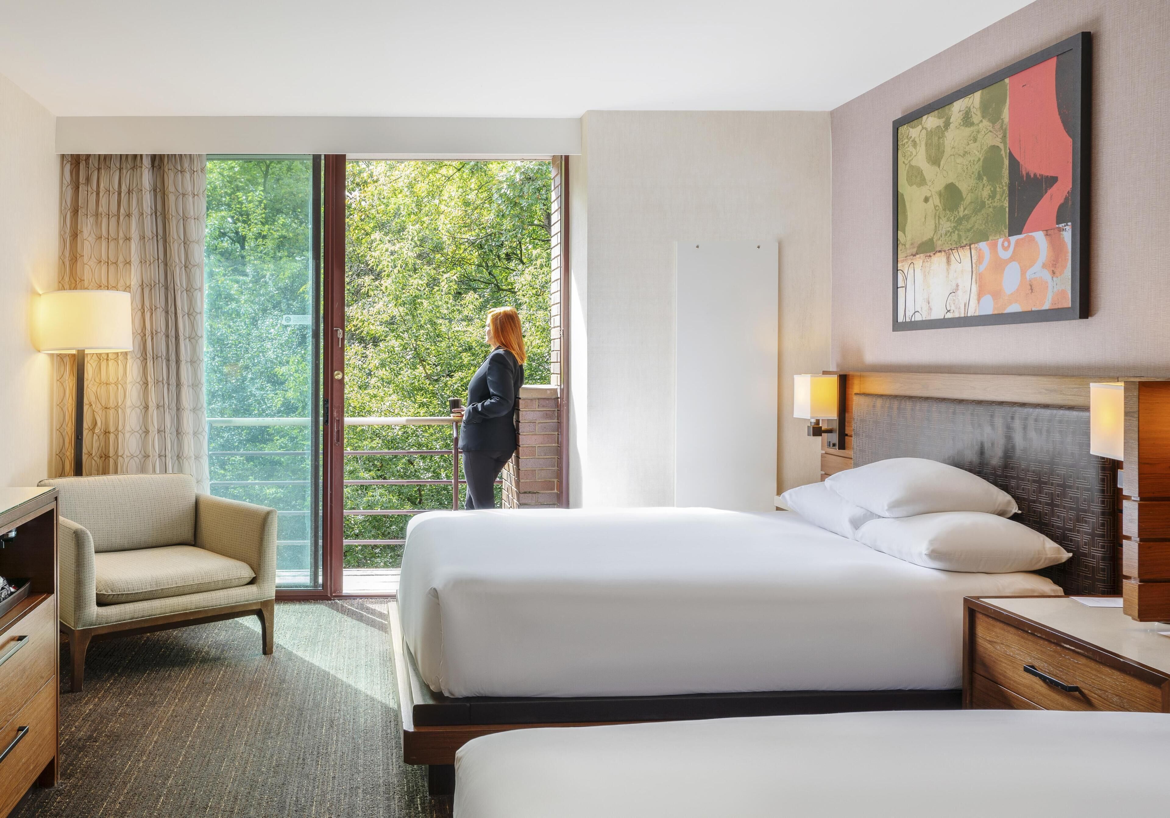 A hotel room with a bed, a chair, a desk, a lamp, a painting, and a balcony with a view of trees.