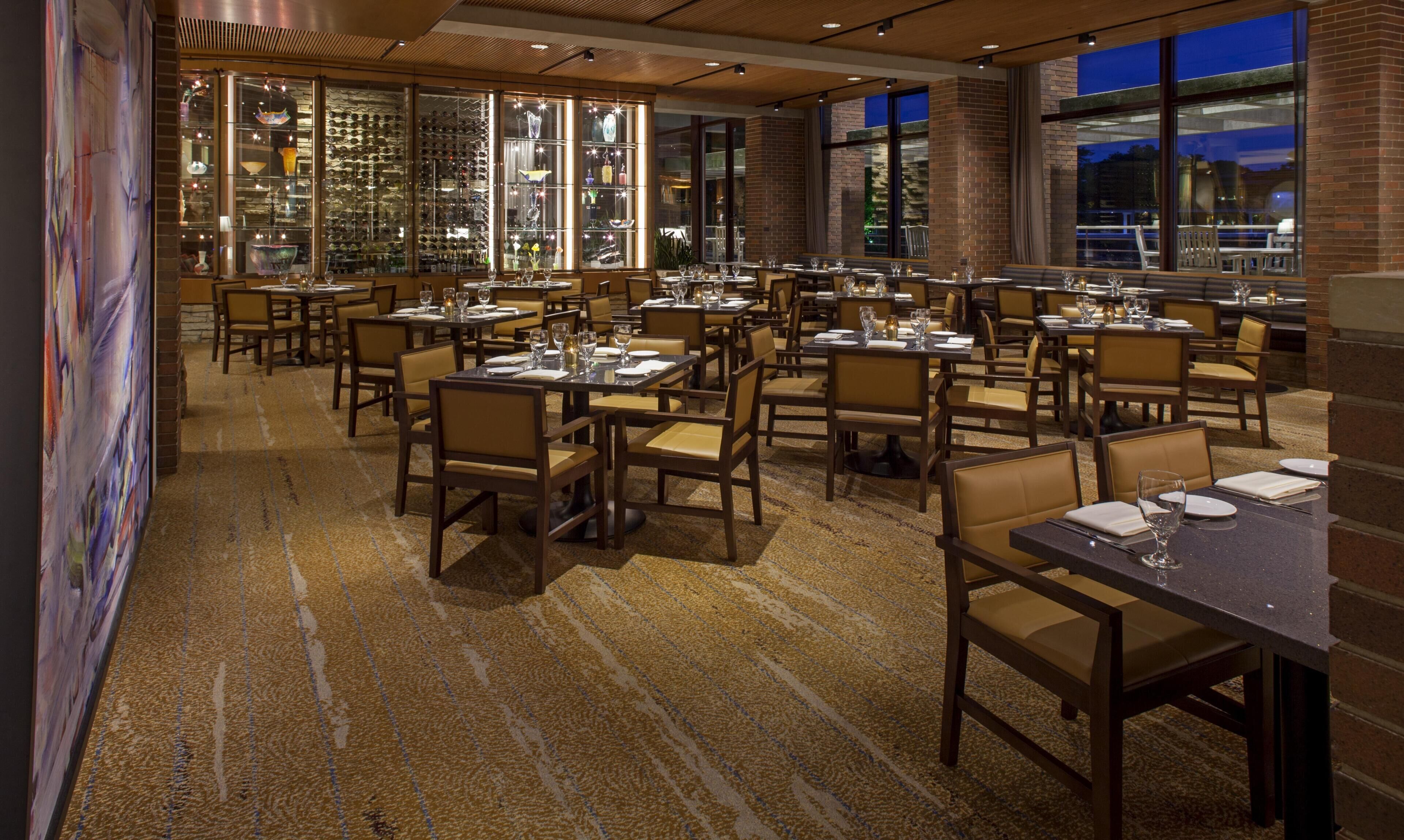 A restaurant with multiple tables and chairs, large windows, and a patterned carpet.