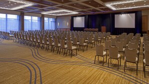 Un salón de conferencias con filas de sillas, un escenario con una pantalla y ventanas amplias.
