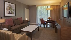Una habitación de hotel moderna con sofá, una mesa de comedor con sillas, televisor y un gran ventanal con cortinas.