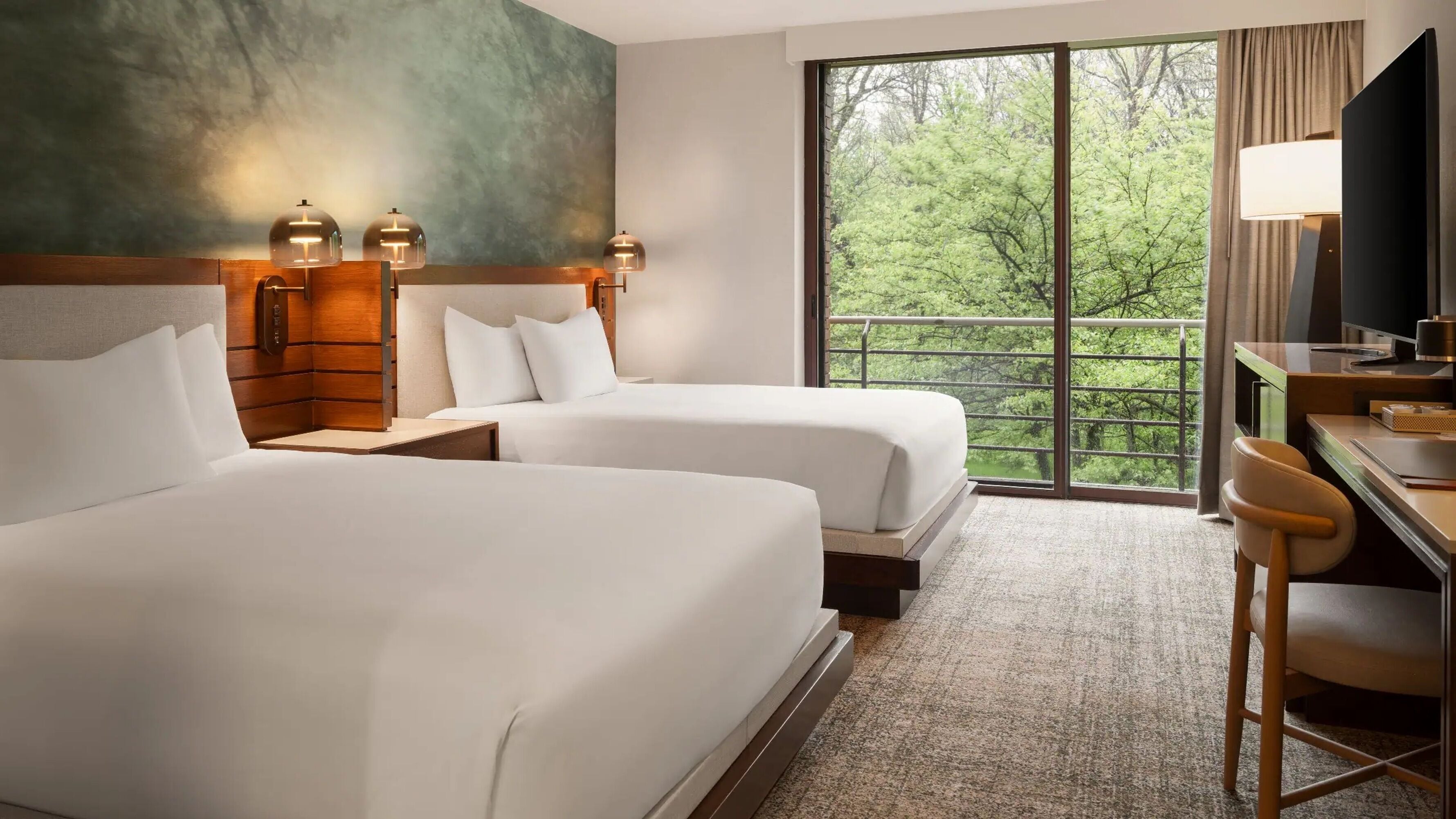 A hotel room with two beds, a desk, a chair, a TV, and a large window with a view of trees.