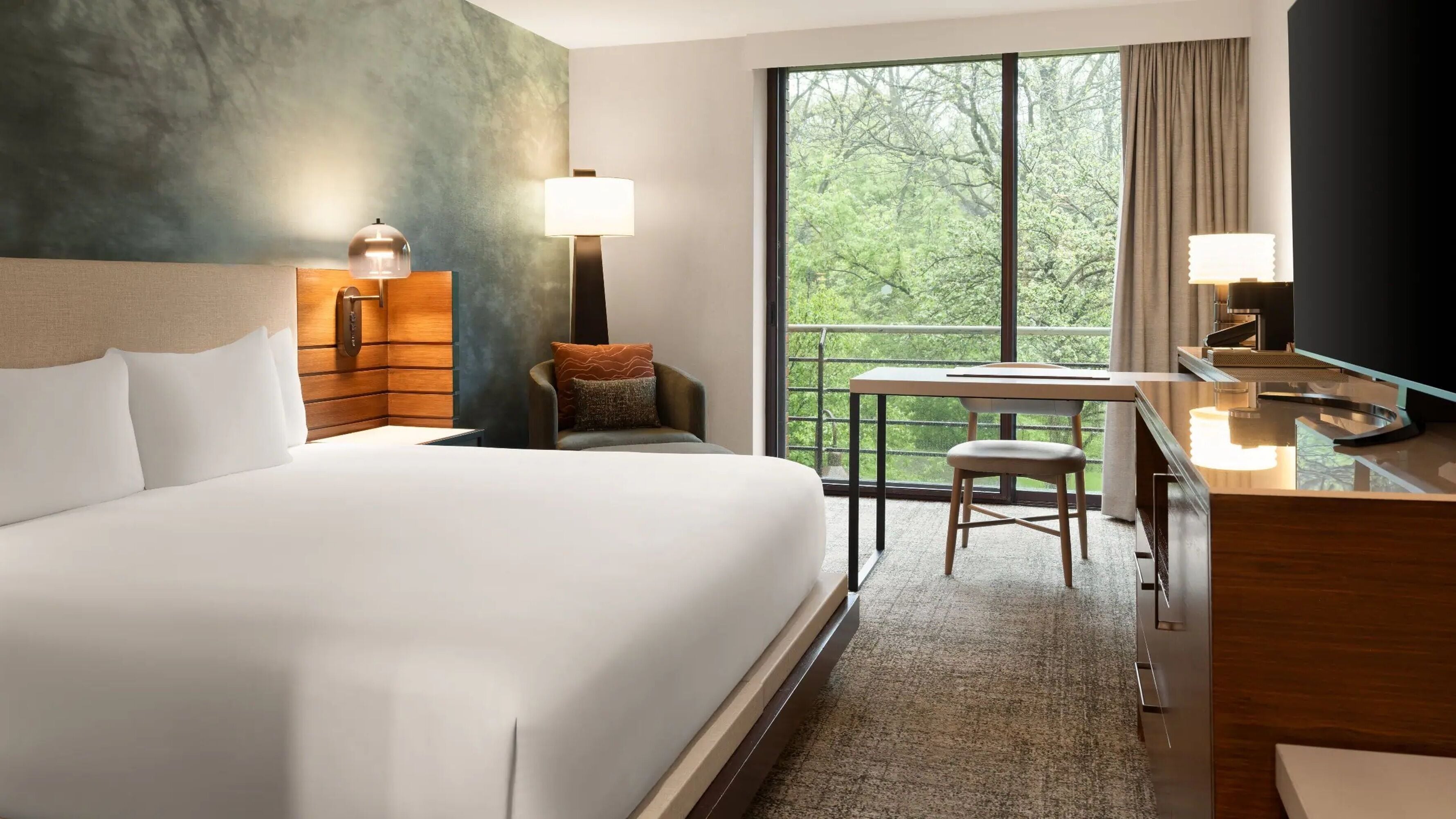 A hotel room with a large bed, a desk, a chair, a lamp, and a view of trees through the window.