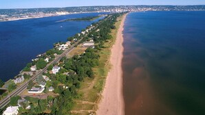 An aerial view of a coastal area with a long stretch of beach, a road running parallel to the shore, and a dense cluster of houses.