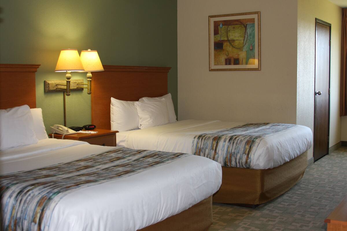A hotel room with two beds, a nightstand with a lamp, a painting on the wall, and a door.