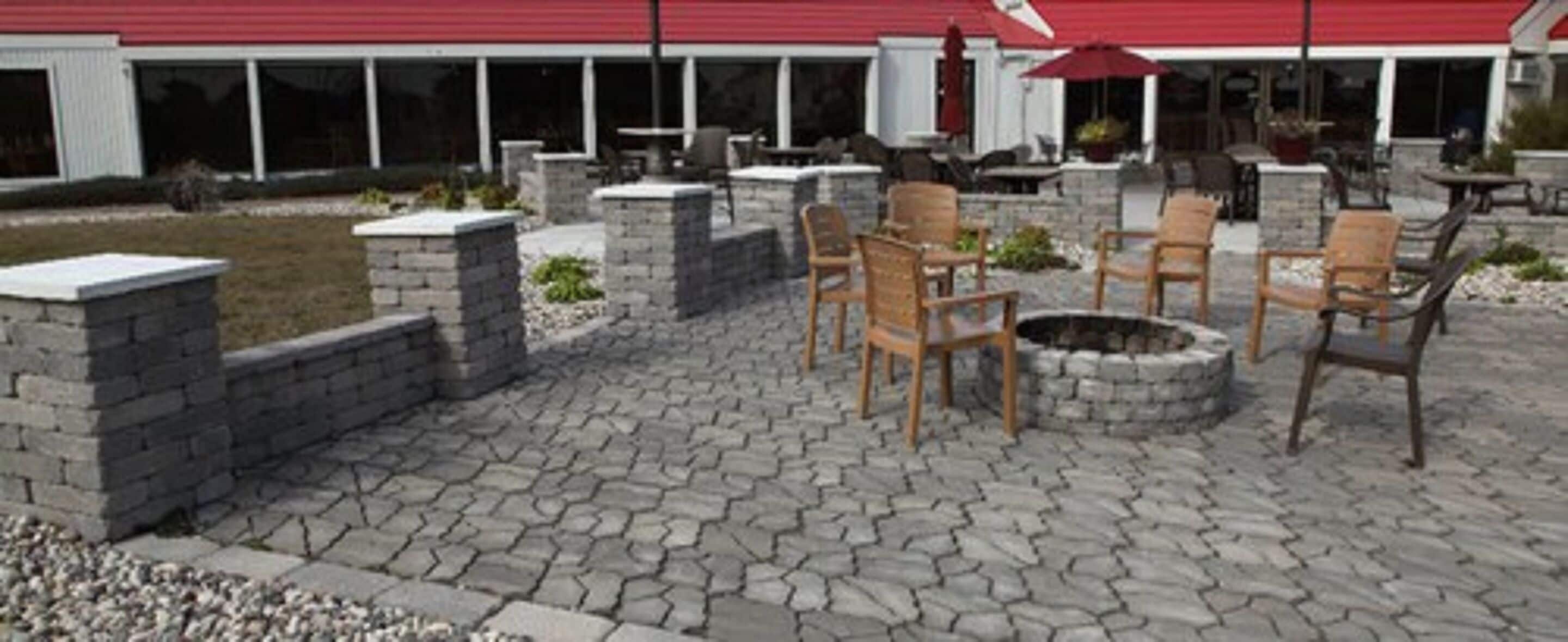 An outdoor patio with stone flooring, wooden chairs, a fire pit, and a building with a red roof in the background.