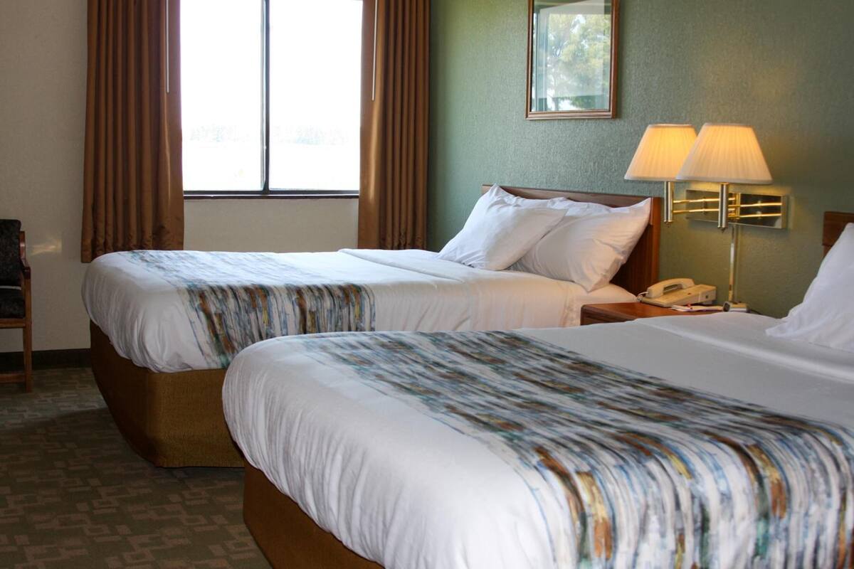A hotel room with two beds, a nightstand with a lamp, a telephone, and a window with curtains.