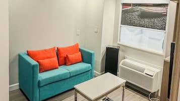Una habitación con un sofá color turquesa, dos cojines anaranjados, una mesa blanca y una ventana con vista a la playa.