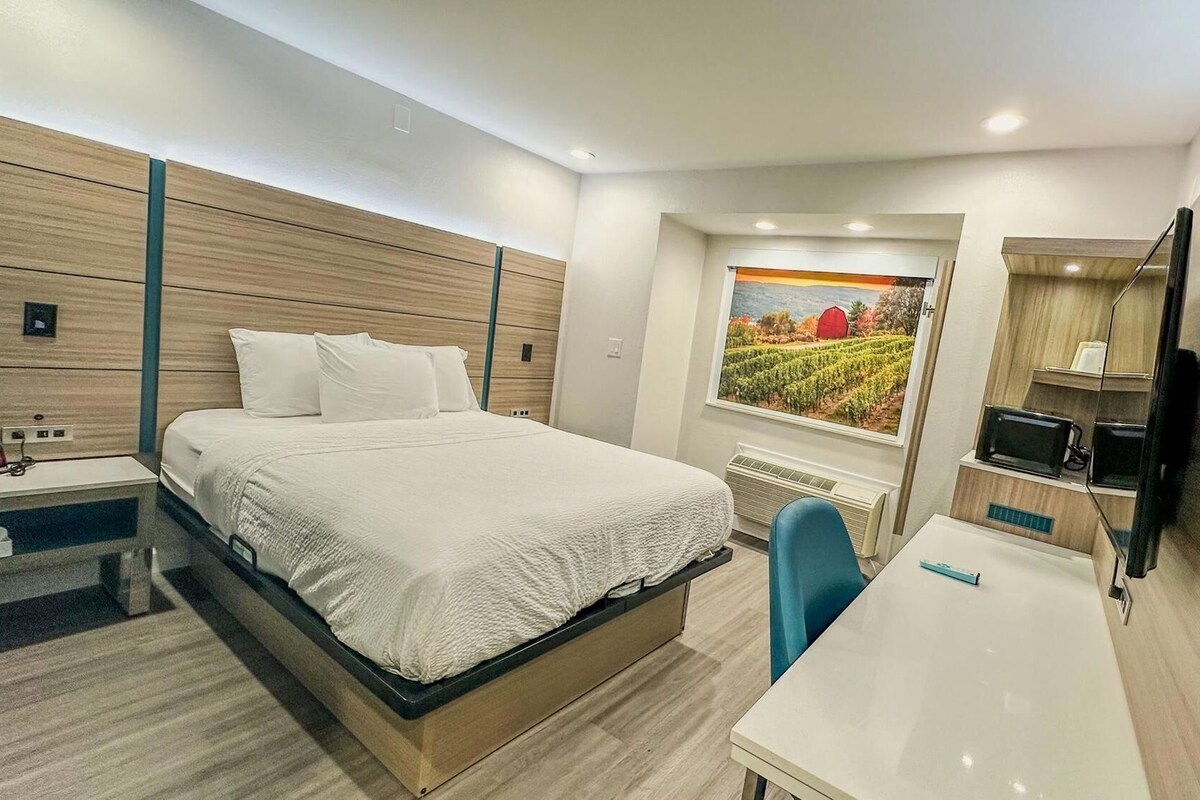 A modern hotel room with a bed, a desk, a chair, a TV, and a painting on the wall.