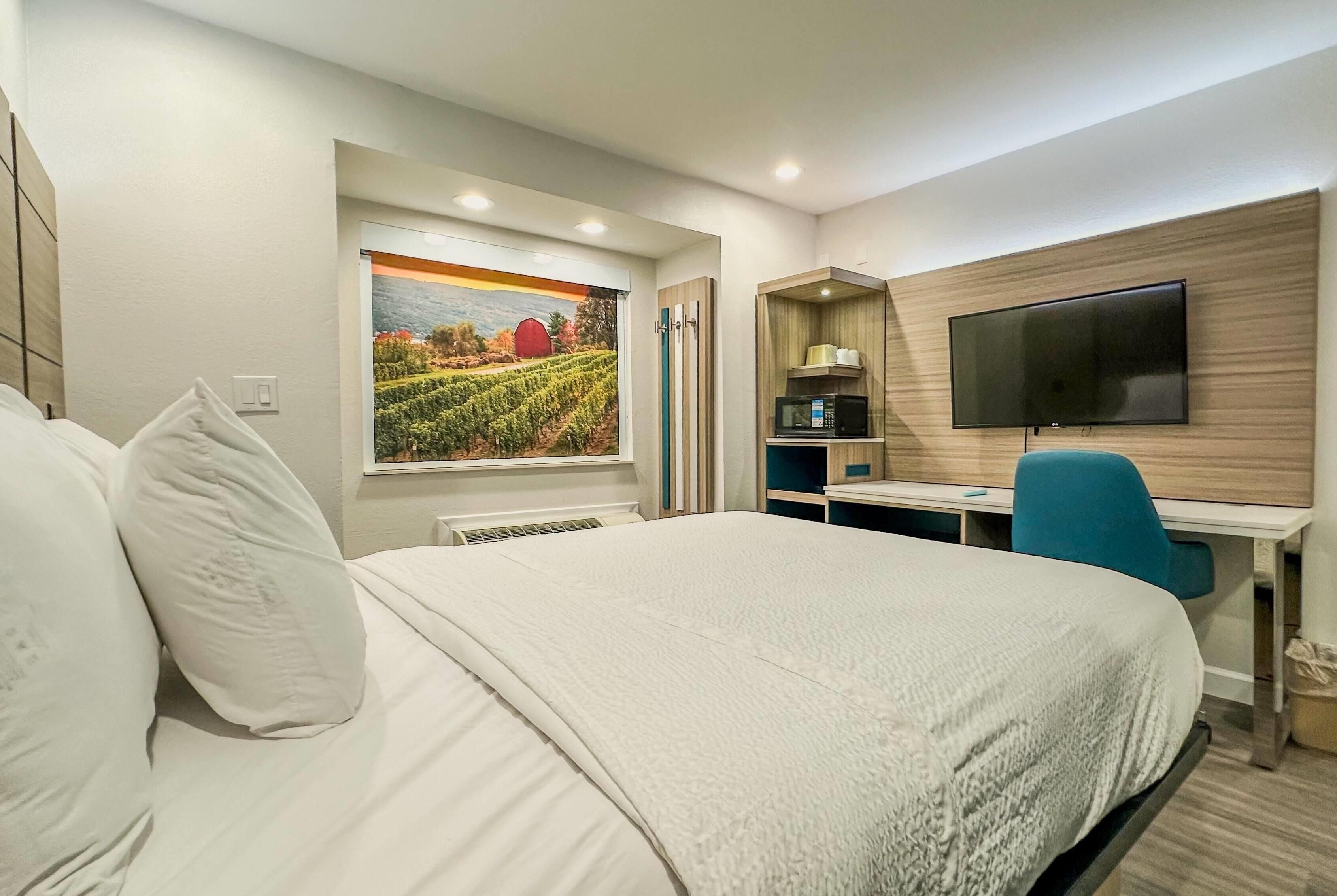 A hotel room with a large bed, a desk with a chair, a flat-screen TV mounted on the wall, and a window with a painting of a vineyard.