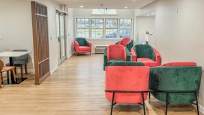 A modern interior with a wooden floor, a dining area with a table and chairs, and a seating area with red and green upholstered chairs. - Baymont by Wyndham Henrietta/Rochester (Henrietta)