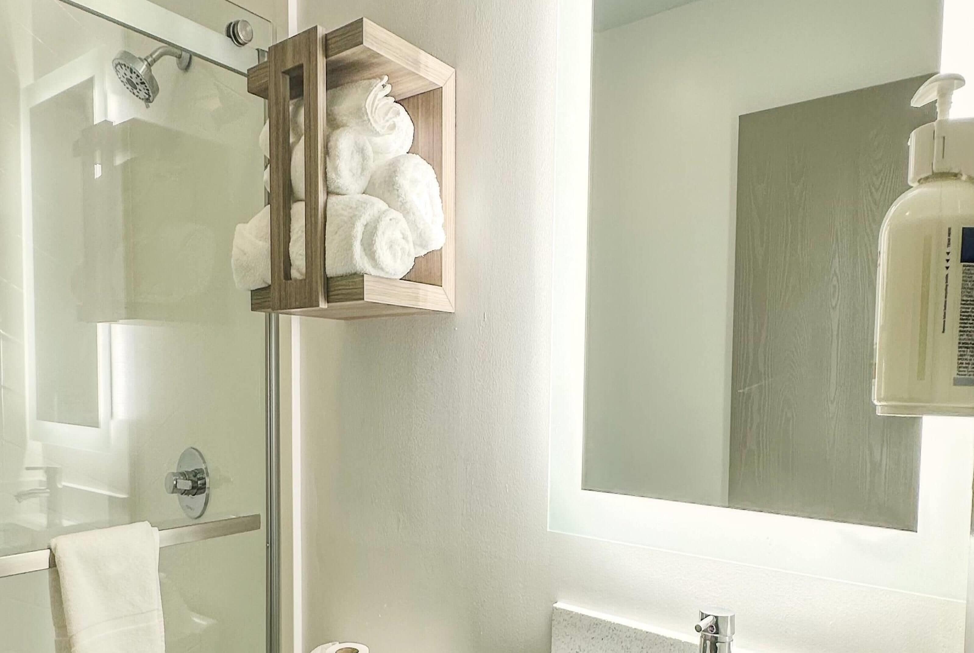 Un baño con cabina de ducha de vidrio, un perchero de madera con toallas blancas, un espejo grande y una botella de jabón para manos.