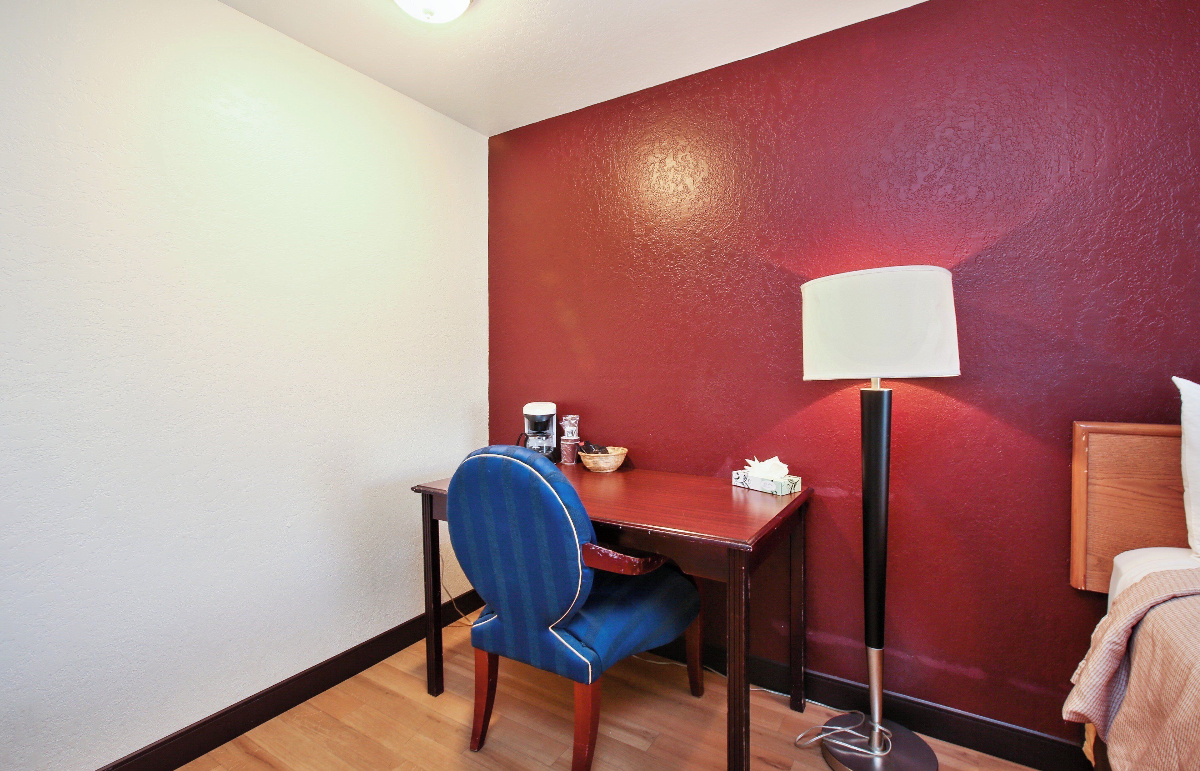 A hotel room with a desk, chair, lamp, and a red wall.