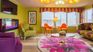 A living room with a flat-screen TV, green armchair, orange chairs, a colorful sofa, and a round coffee table.