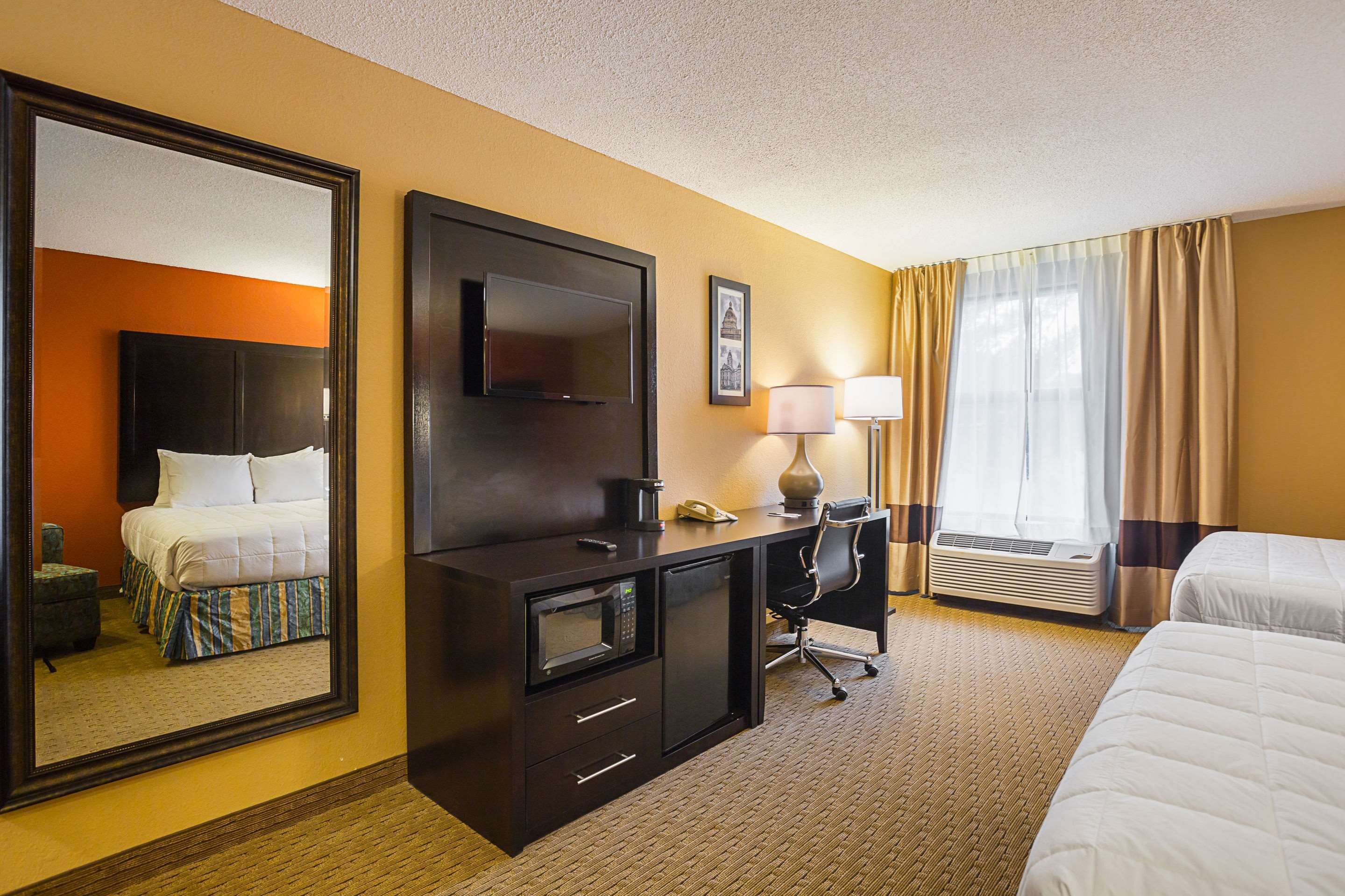 Habitación de hotel con cama, escritorio, computadora, televisor y un espejo grande.