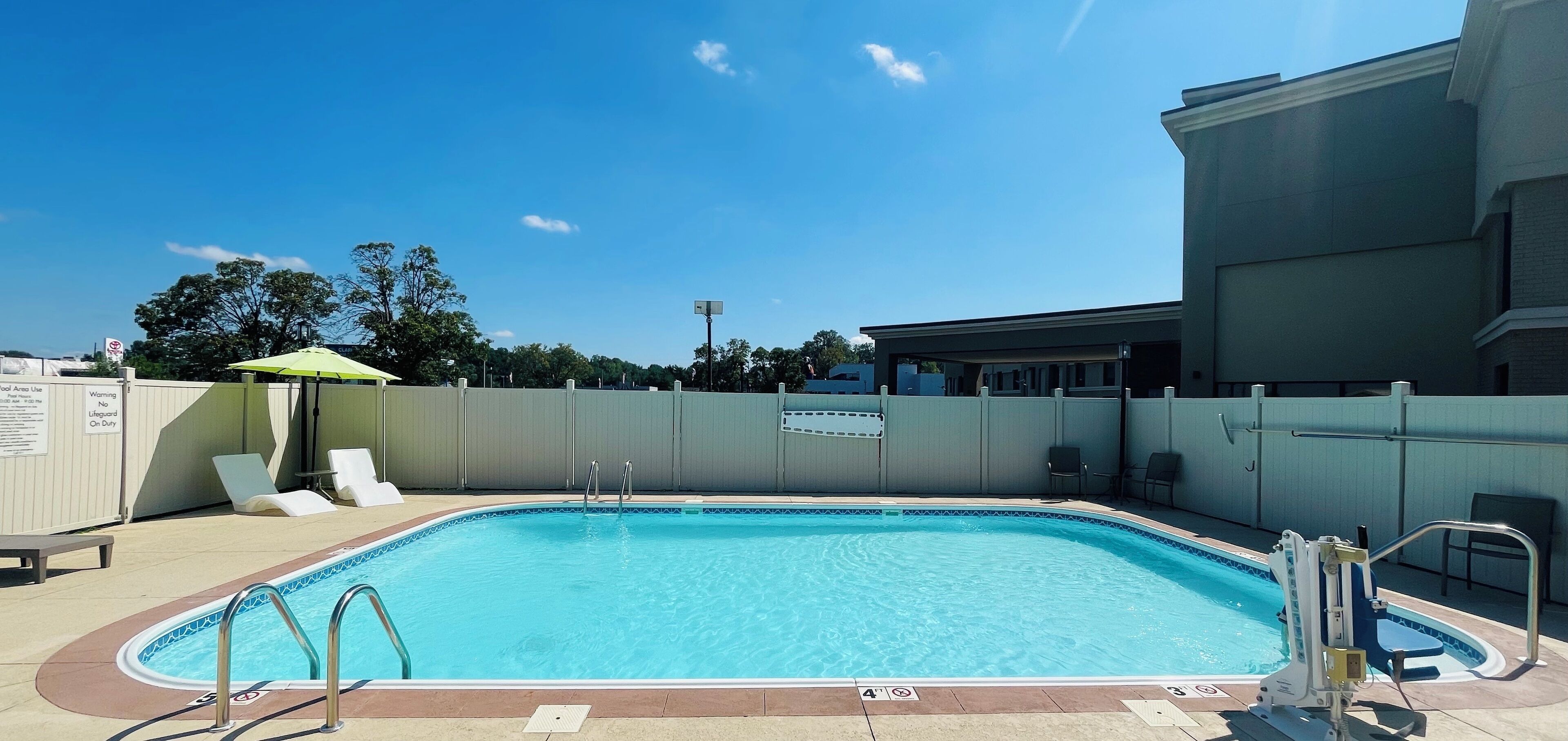 Ein rechteckiger Pool mit Fliesenrand, umgeben von einer niedrigen Mauer und einem Metallzaun. In der Nähe befinden sich Liegestühle und ein grüner Sonnenschirm.
