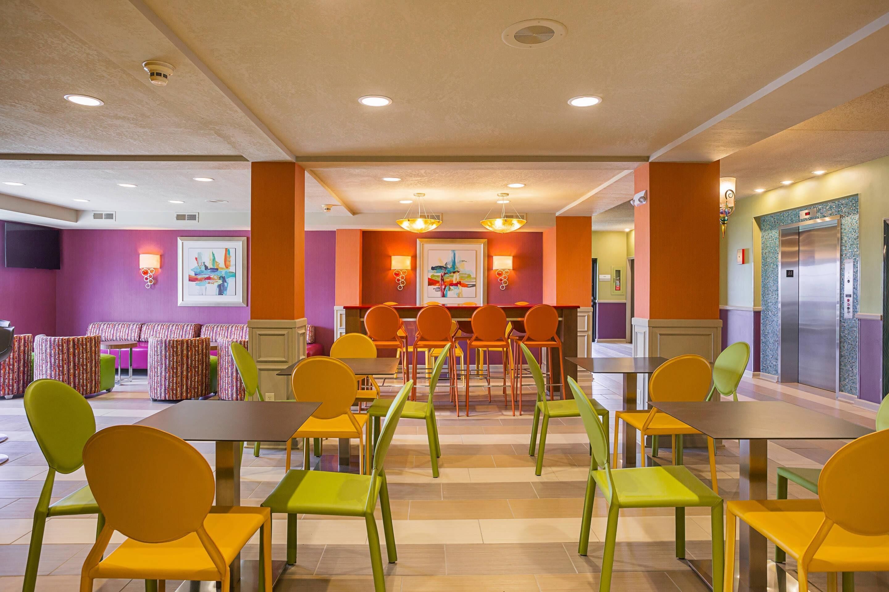 Un comedor bien iluminado con sillas y mesas coloridas, una zona de estar con bancos acolchados y un ascensor visible.