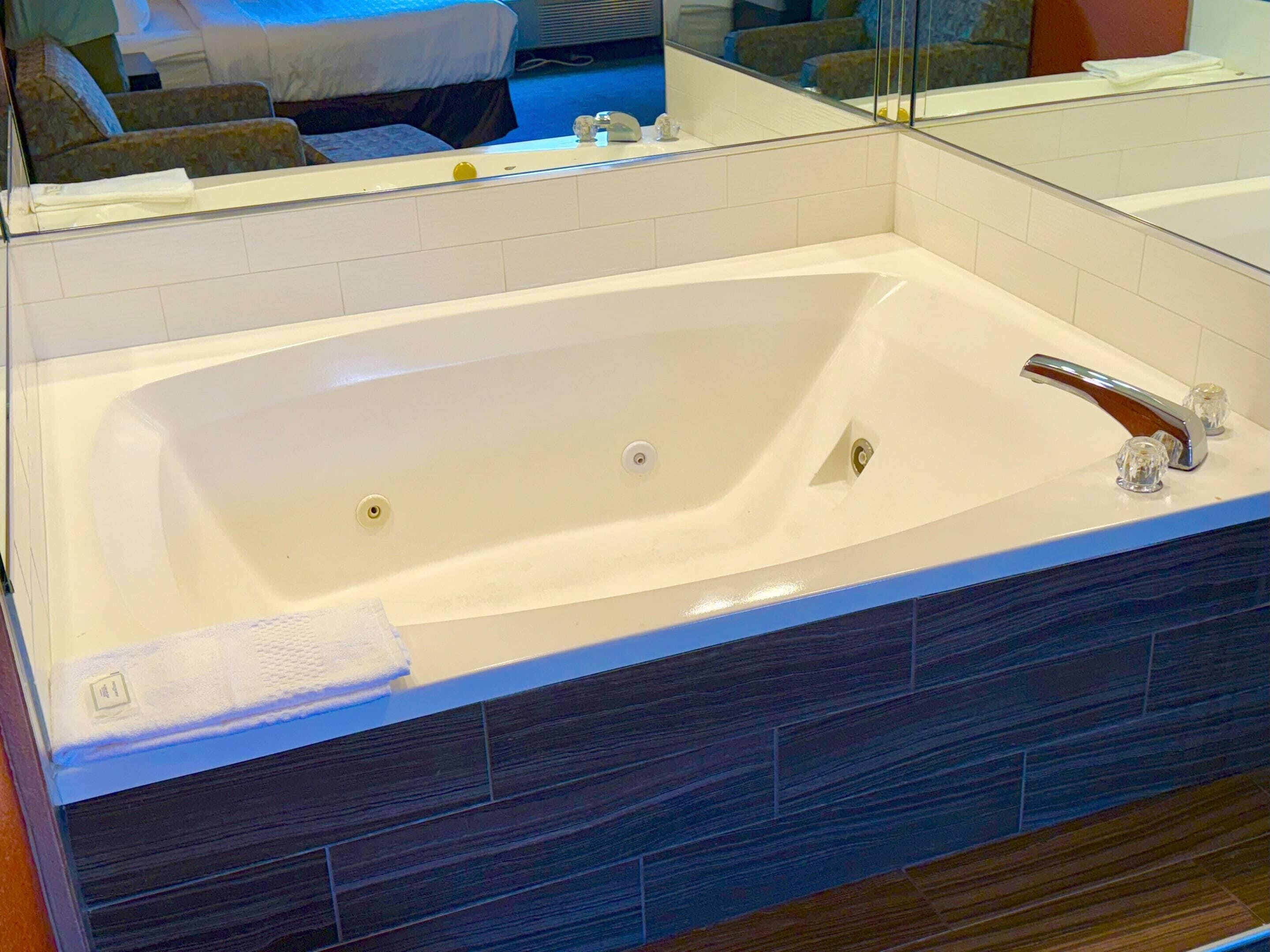 A modern bathroom with a large bathtub, a glass shower enclosure, and a visible bed in the reflection.