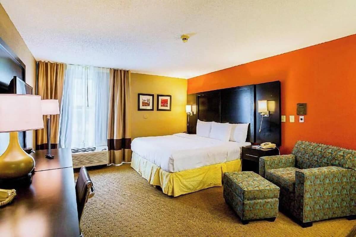 Una habitación de hotel con cama, escritorio con lámpara, televisor, sofá y reposapiés.