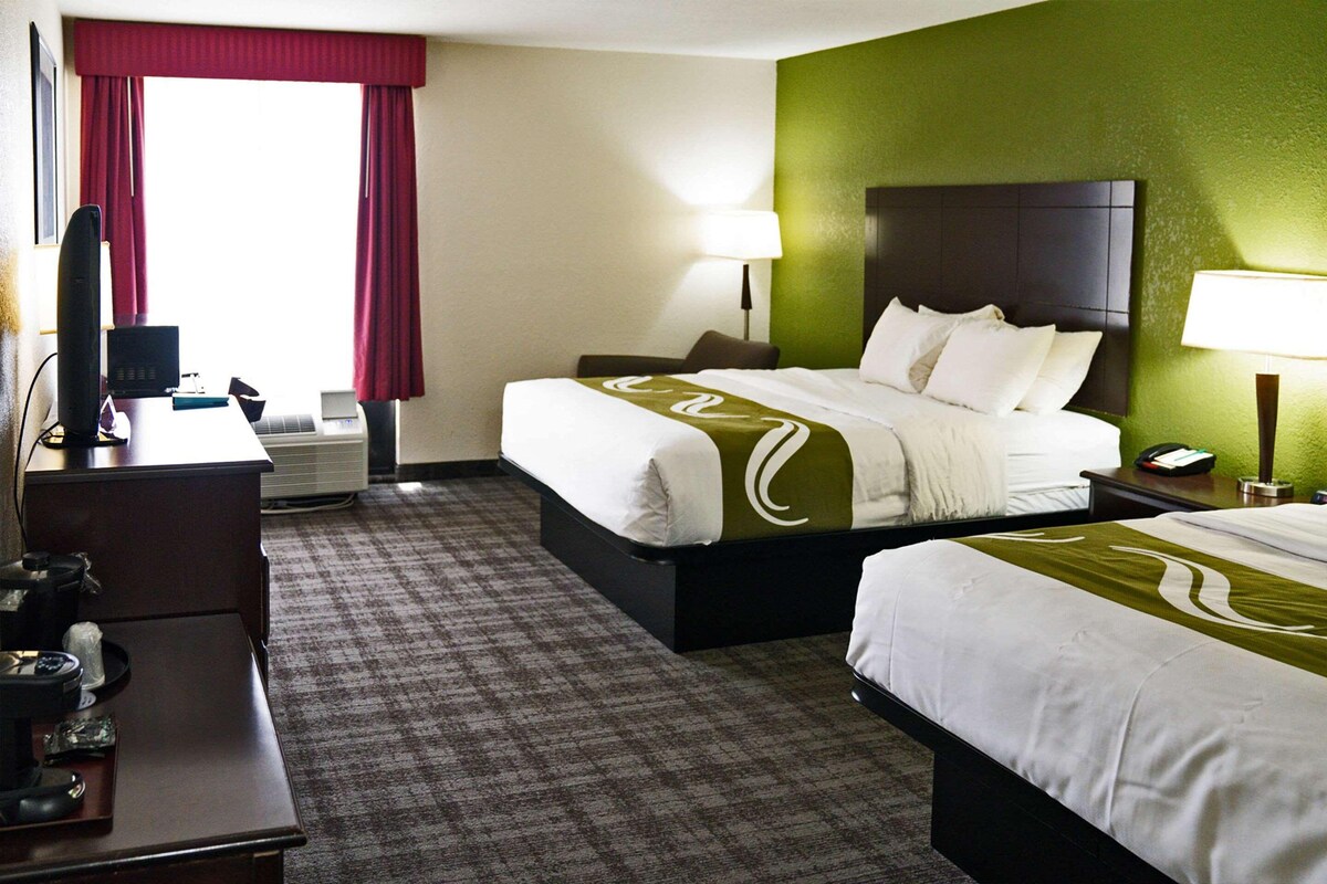 A hotel room with two beds, a desk, a chair, a TV, and a lamp.
