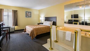 A hotel room with a bed, a desk, a chair, a large mirror, and a bathtub. - Quality Inn & Suites Greensburg I-74 (Greensburg)