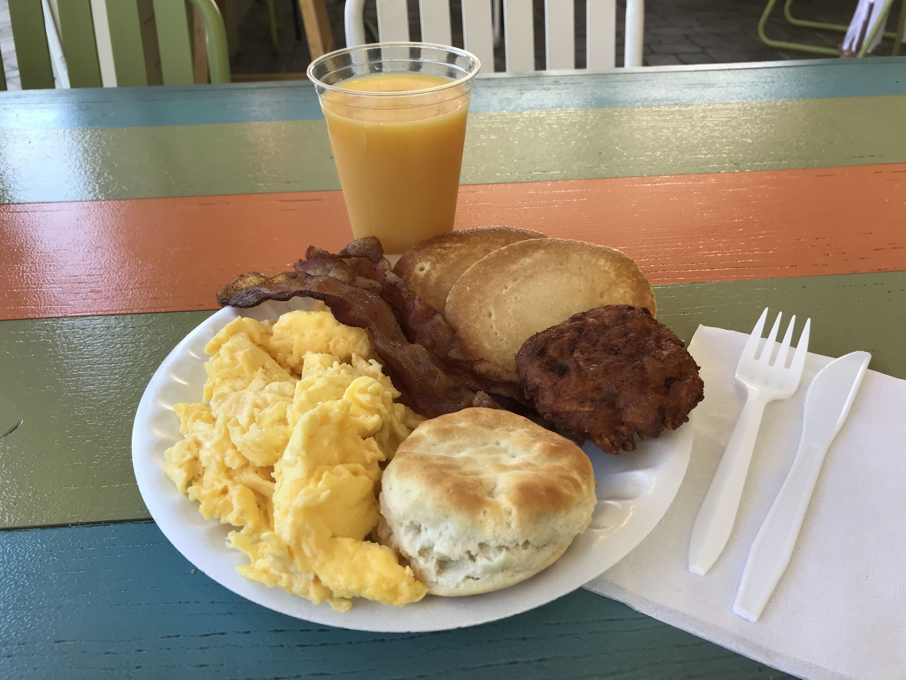 A plate with scrambled eggs, bacon, sausage, a biscuit, and a glass of orange juice on a colorful table.