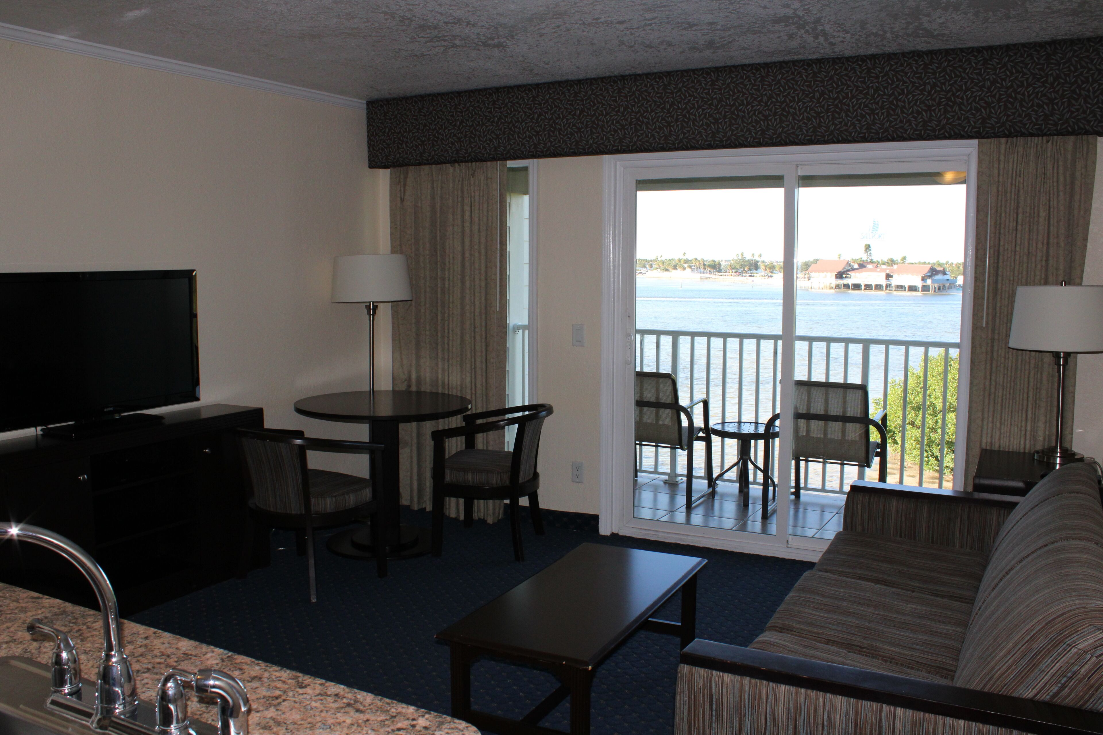 Une chambre d’hôtel équipée d’une télévision, d’une table à manger avec des chaises, d’un canapé, d’un balcon avec vue sur l’eau et d’une grande fenêtre.