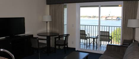 Une chambre d’hôtel équipée d’une télévision, d’une table à manger avec des chaises, d’un canapé, d’un balcon avec vue sur l’eau et d’une grande fenêtre.
