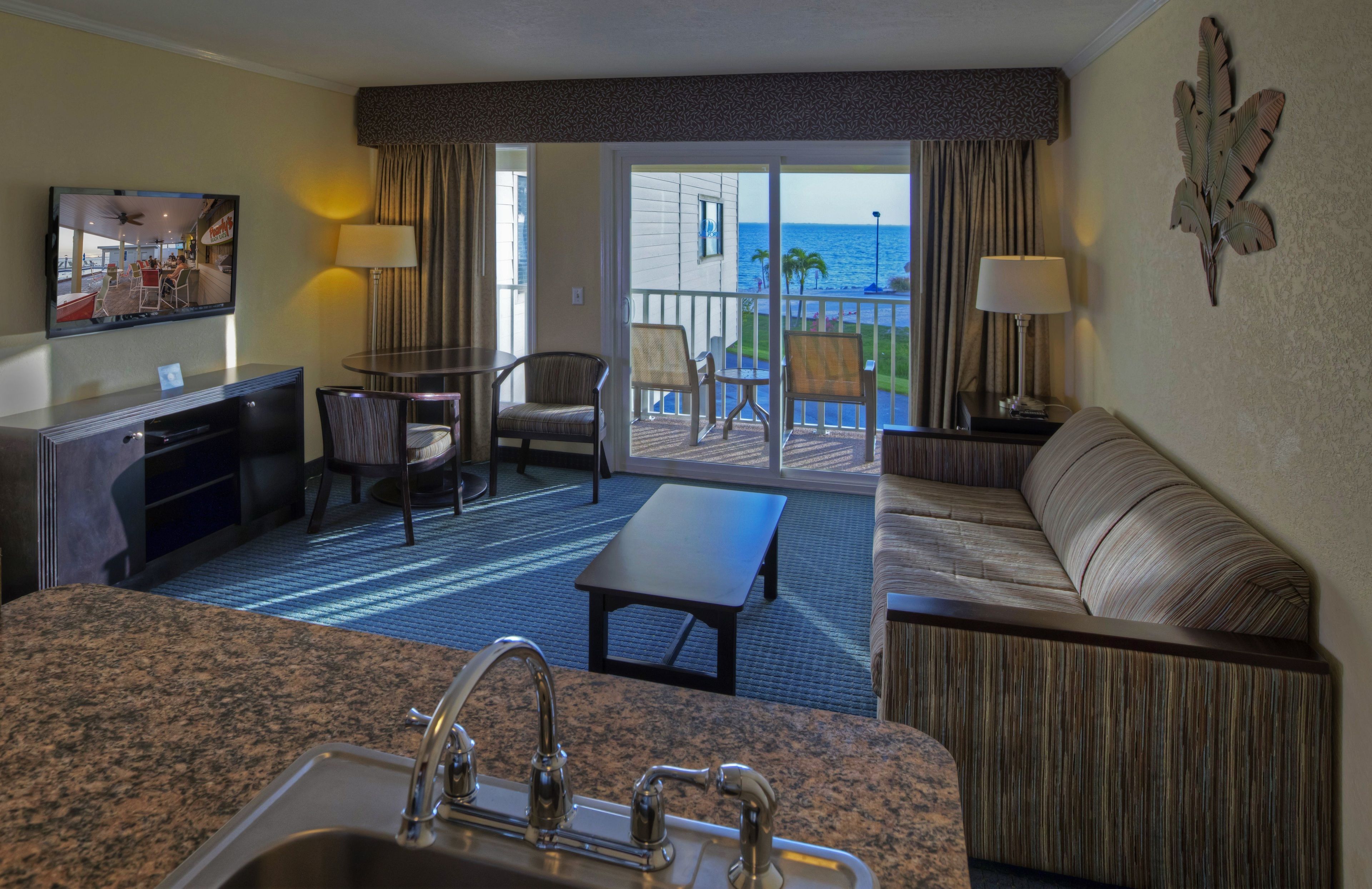 A hotel room with a kitchen area, a seating area with a sofa and chairs, a balcony with a view of the ocean, and a television mounted on the wall.