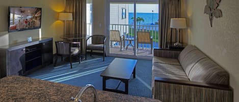 A hotel room with a kitchen area, a seating area with a sofa and chairs, a balcony with a view of the ocean, and a television mounted on the wall.