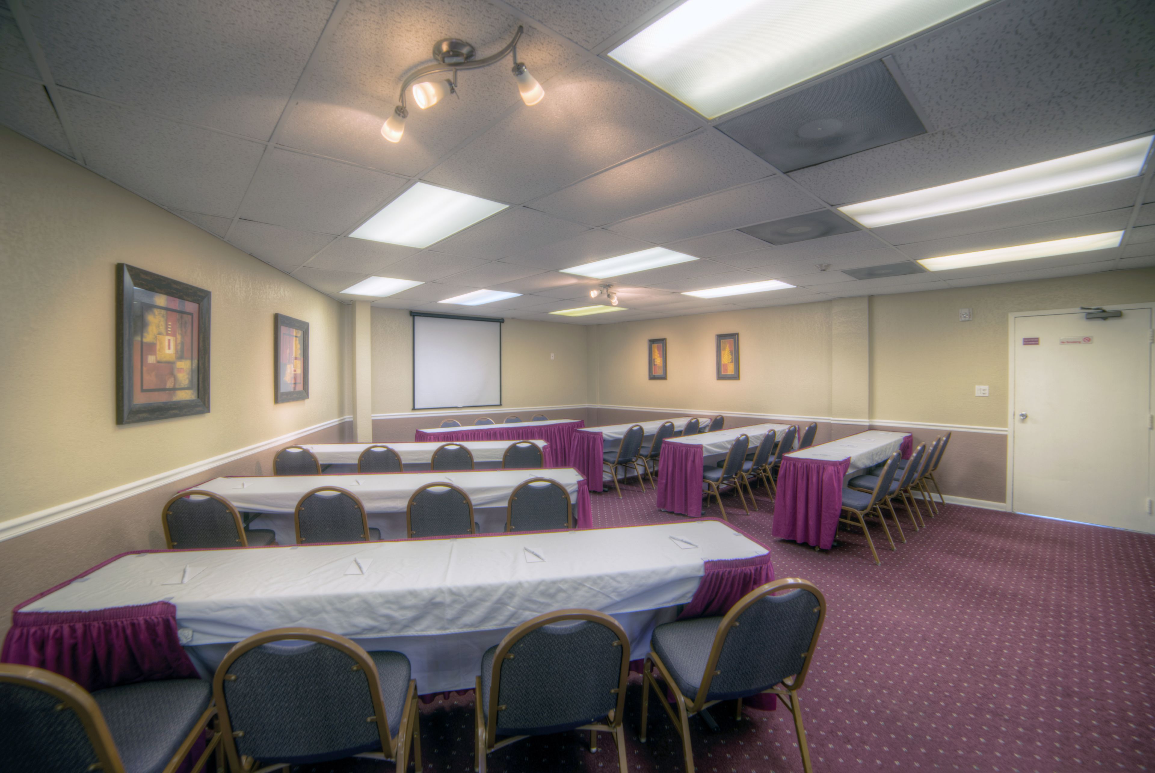 A conference room with tables and chairs, a projector screen, and framed pictures on the walls.