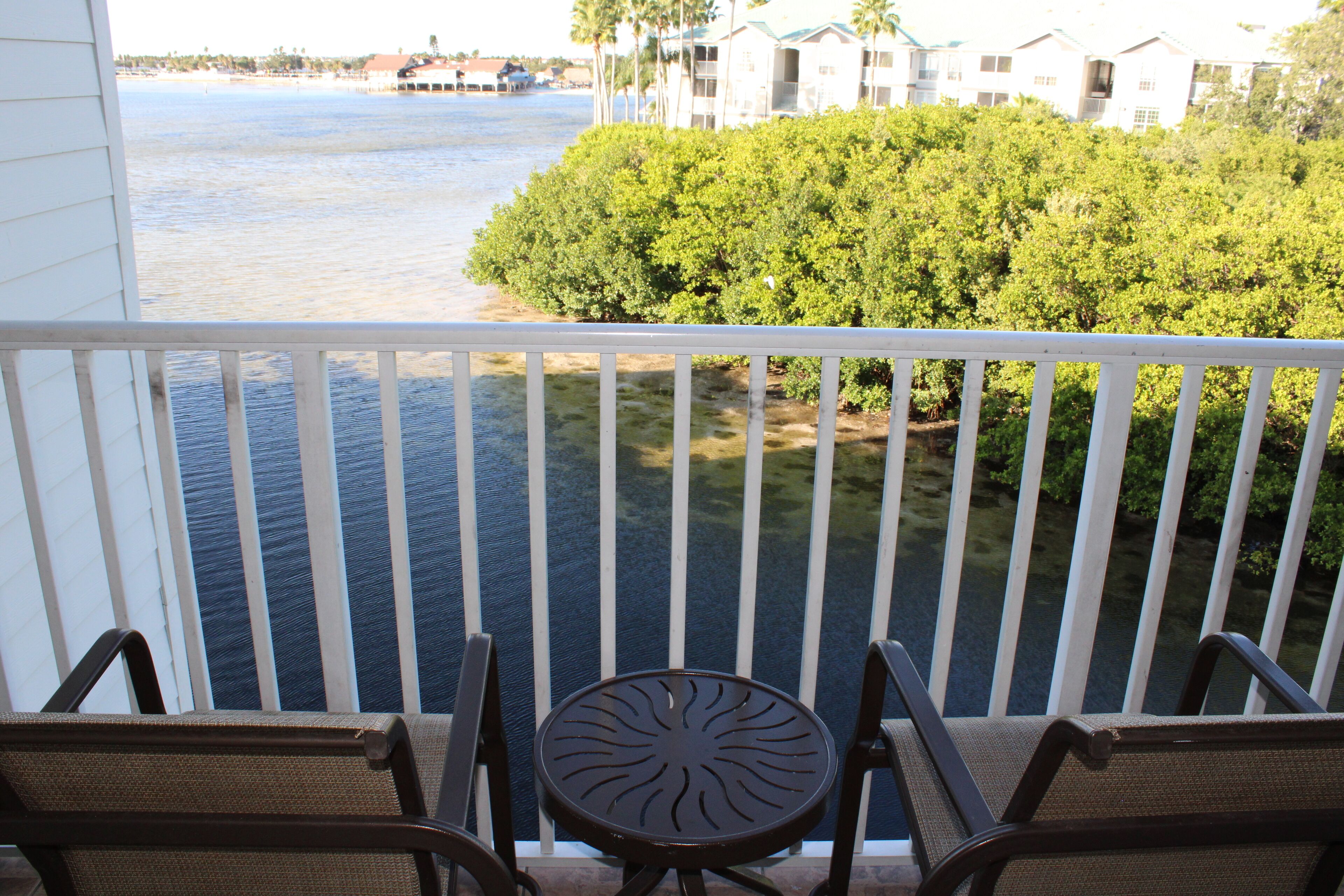A balcony with two chairs and a table, overlooking a body of water and a green bush.