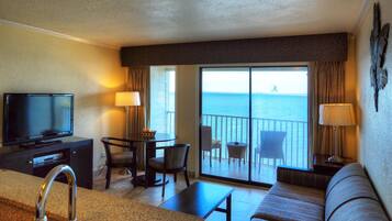 A hotel room with a TV, a sofa, a dining table with chairs, a balcony with a view of the sea, and a large window.