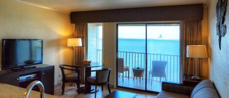 Habitación de hotel con televisor, sofá, mesa de comedor con sillas, balcón con vistas al mar y un ventanal grande.