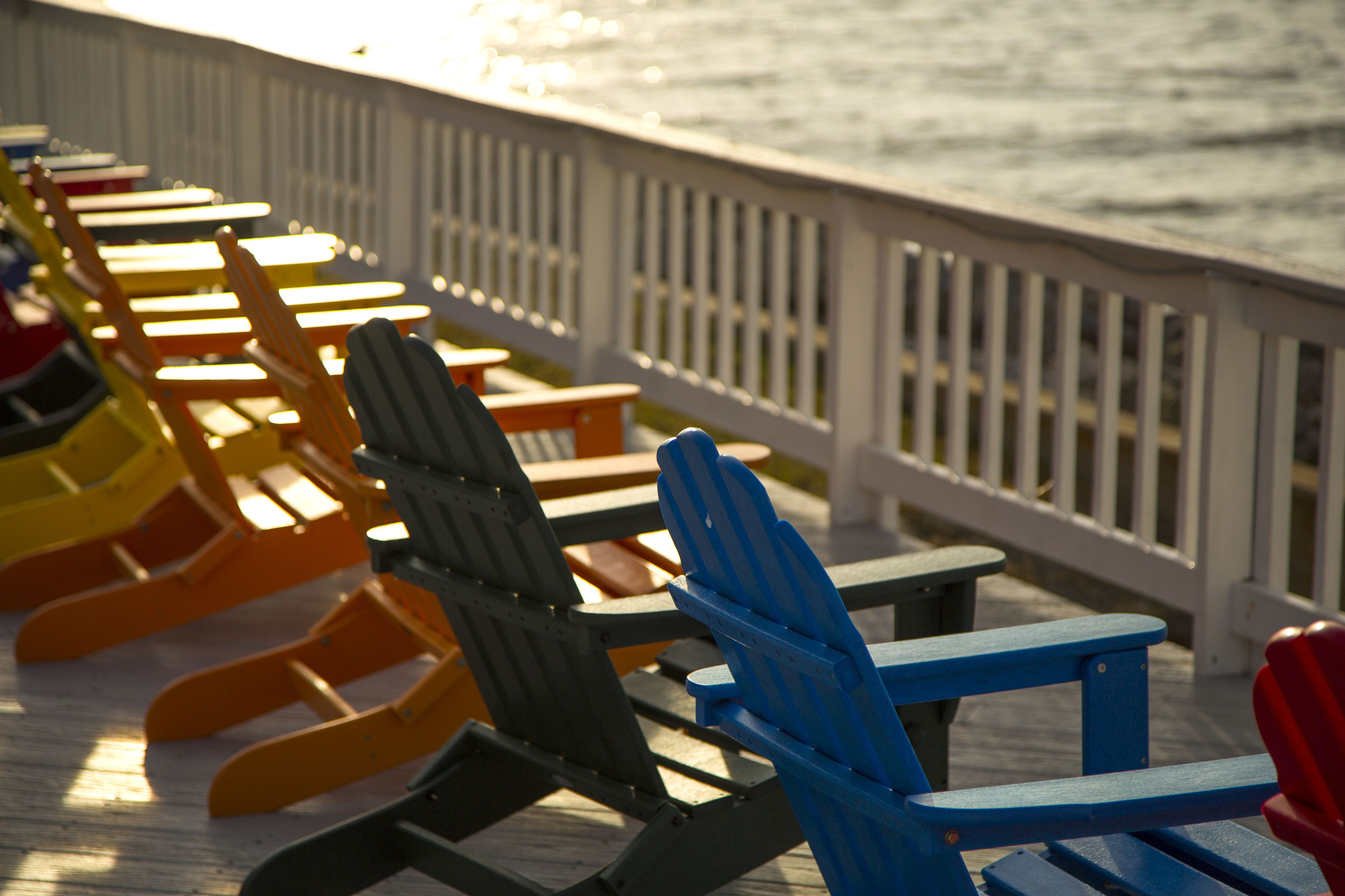 Une rangée de chaises Adirondack aux couleurs vives est disposée sur une terrasse en bois dotée d’une balustrade, et elle offre une vue sur une étendue d’eau.