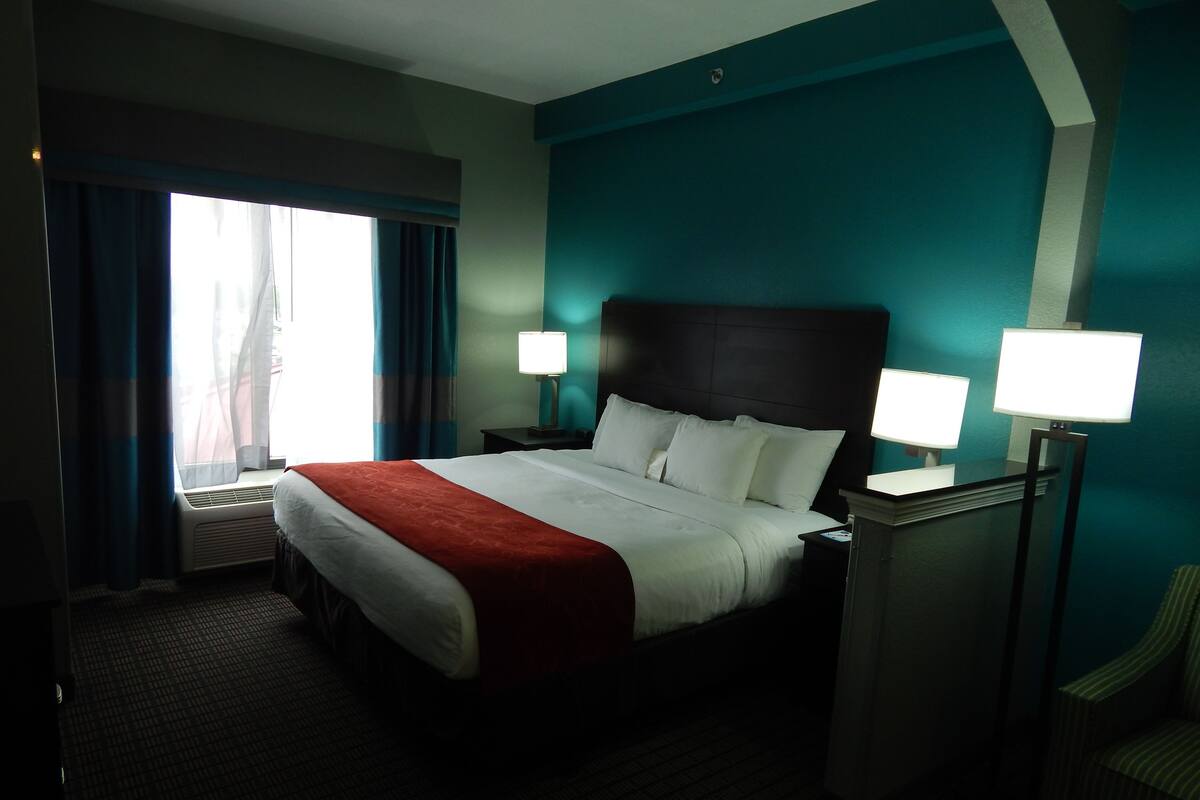 Une chambre d’hôtel avec un grand lit, deux lampes de chevet, une fenêtre avec des rideaux et un mur d’accent de couleur turquoise.