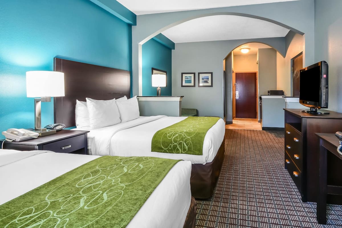 Une chambre d’hôtel avec deux lits, un bureau avec une lampe, une télévision et une salle de bain visible à travers une arche.
