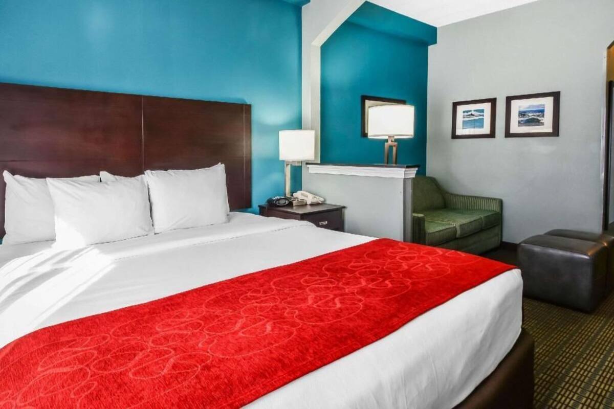 Une chambre d’hôtel avec un grand lit, une table de chevet, une lampe, un fauteuil vert et un pouf gris.