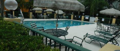 A poolside area with lounge chairs and umbrellas, surrounded by greenery and a building in the background.