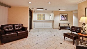 A spacious living area with a brown leather sofa, a wooden coffee table, and a chair. A small table with a lamp is also visible. The room has a tiled floor and a white cabinet in the background. - La Quinta Inn & Suites by Wyndham Tampa Fairgrounds - Casino (Tampa)