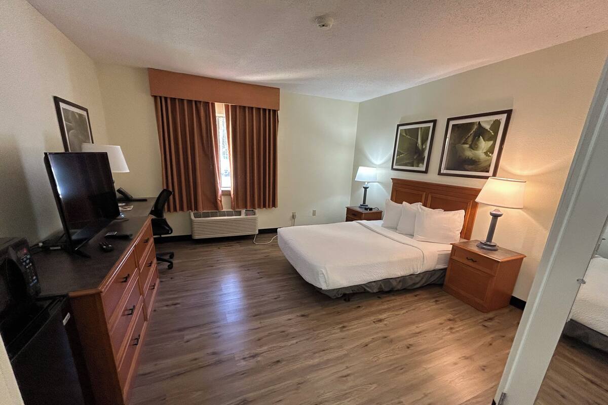A hotel room with a bed, a desk with a computer, a chair, a dresser, a lamp, and a window with curtains.