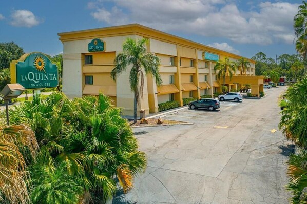 A hotel with a sign reading "La Quinta Inn & Suites," featuring a multi-story building with a yellow facade, palm trees, and a parking lot with several cars.