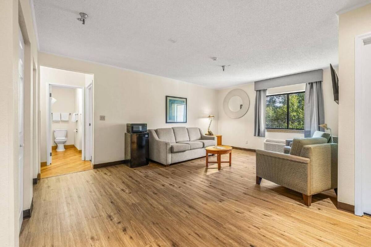 A spacious living room with a wooden floor, a grey sofa, a small round table, and a wicker chair. A window with curtains is visible, and a door leads to a bathroom.